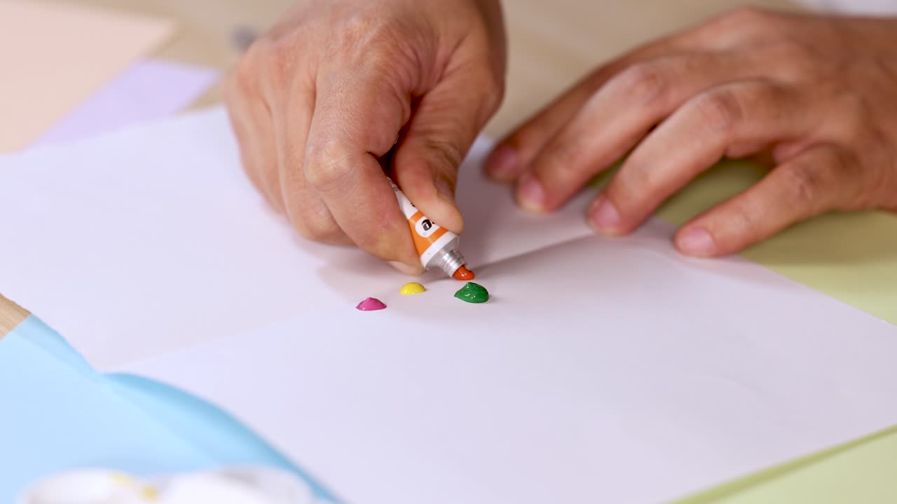 Hands squeeze paint tubes, dispensing colorful drops onto white paper in bright, soft lighting