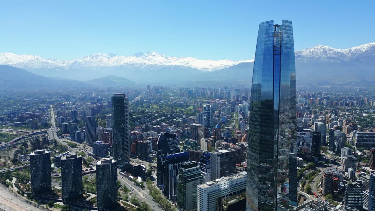 Drone aerial forward approach toward the Costanera skyscrapers and snowcapped Andes, flying over highways, rooftops and treelines in clear daytime light. Santiago, Chile