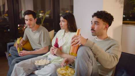 side view of three friends watching interesting movie on television sitting on couch, eating popcorn and chips and drinking soda 2