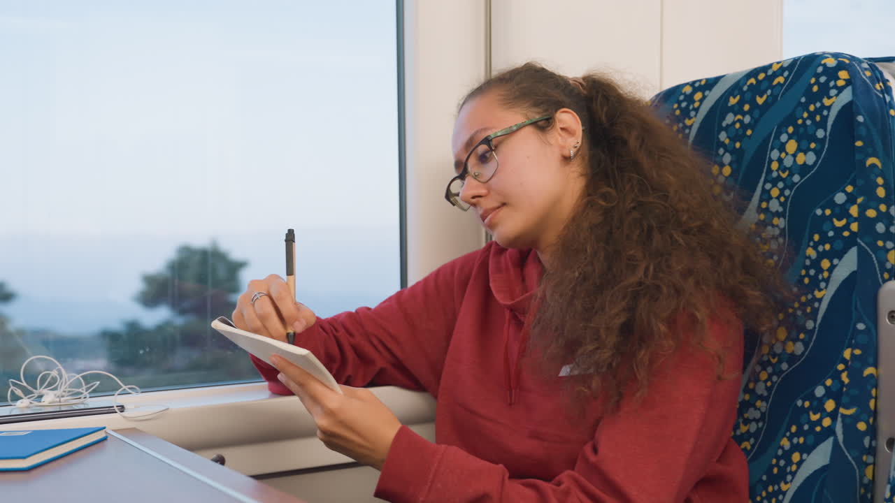 Young woman journaling on train seat, red sweater, curly hair and glasses, writing in notebook beside window, passing landscape and soft light, focused student sketching study notes, travel planner