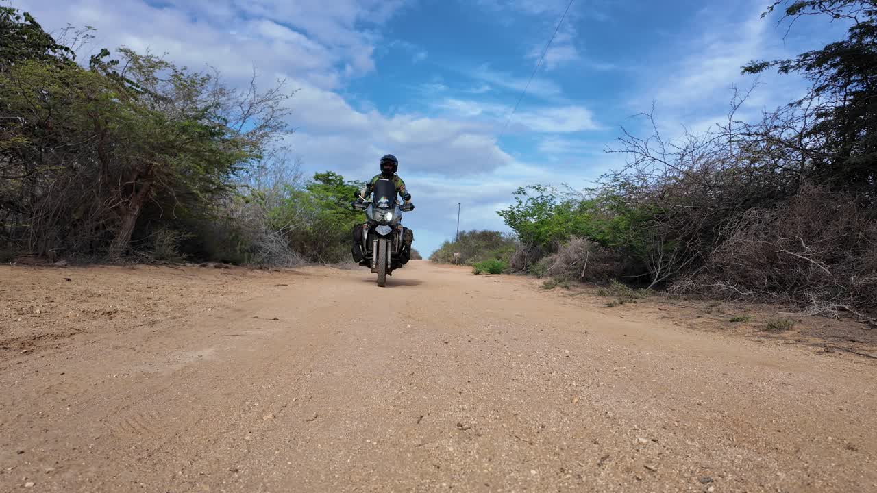 Low-angle, static shot of a fully equipped dual-sport motorcyclist riding directly toward the camera on a dusty, rugged path in a dry, scrubland environment