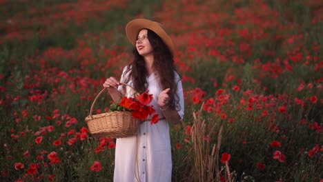 Hermosa-Chica-De-Cabello-Oscuro-En-Un-Campo-De-Flores-Silvestres-Y-Amapolas-Rojas,-Con-Un-Sombrero-Y-Un-Vestido,-Sosteniendo-Una-Canasta-Tejida-De-Flores-Y-Sonriendo