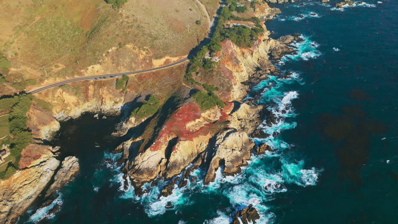 Blue water splashing by the craggy shore of California. Green trees growing along the highway on the rocky coastline. Aerial perspective.