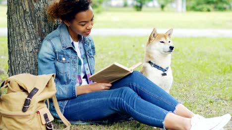 retrato de una bonita estudiante afroamericana leyendo un libro en un parque sentada en la hierba con su obediente perro cerca.