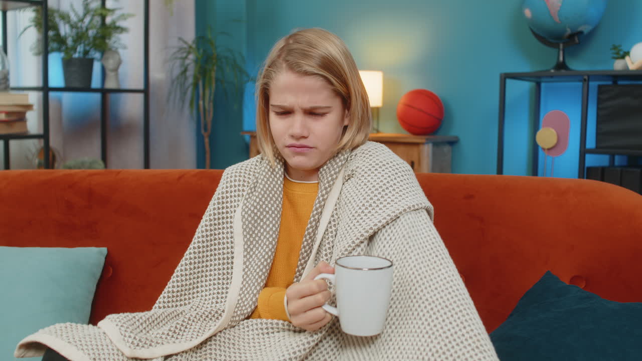 Teenager boy sick with cold and allergy covered with blanket drinking warm tea coughing at home