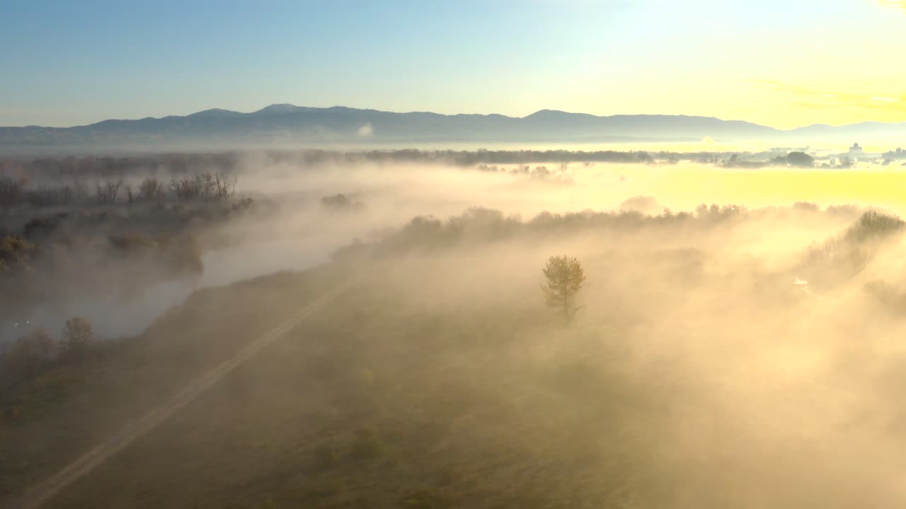 Aerial over farm land and a pond with thick fog during a bright sunrise