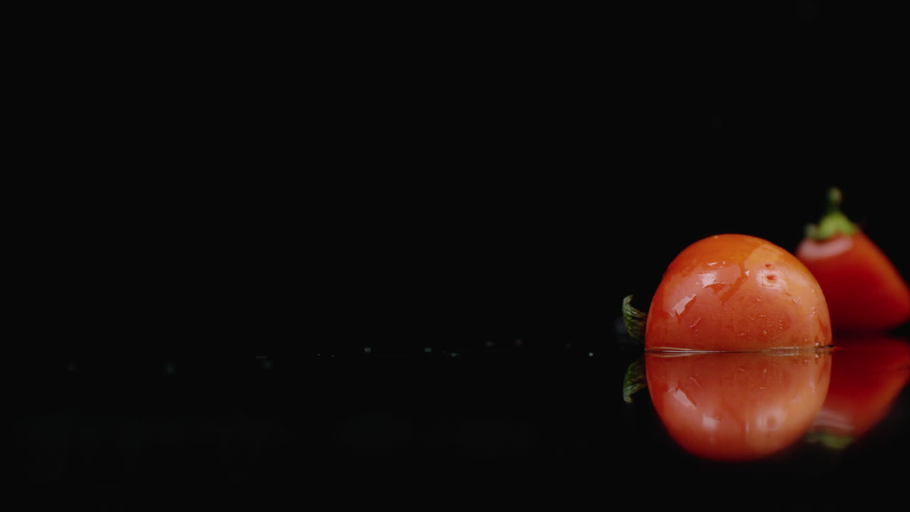빨간 토마토는 어두운 배경에 천천히 움직이는 물의 스프레이와 함께 4 부분의 유리로 떨어집니다.
