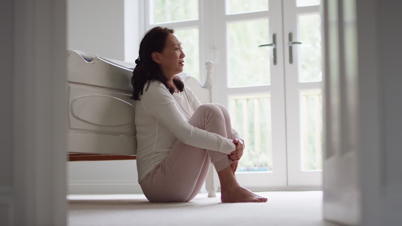침실 바닥에 앉아 있는 피자마를 입은 불행하고 우울한 성숙한 아시아 여성 - 느린 동작으로 촬영