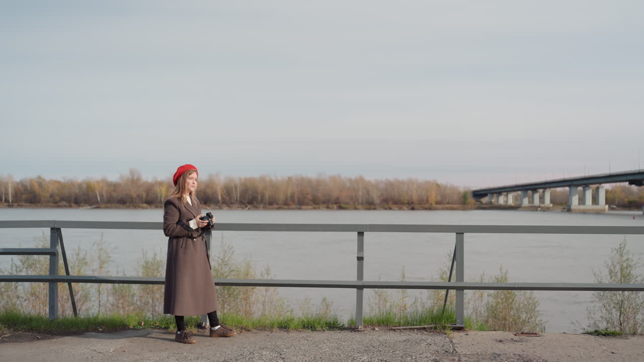 Женщина в пальто и берете у воды. Непринужденная городская сцена с женщиной, проверяющей свой телефон у реки. Женщина в коричневой куртке и красной шляпе изучает свое устройство на берегу реки в городе.