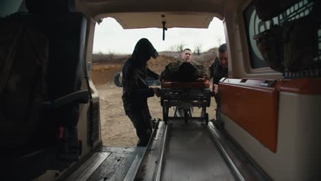 Vista-Desde-El-Costado-De-La-Cabina-De-La-Ambulancia.-Tres-Militares-Colocan-A-Un-Hombre-Herido-Con-Uniforme-De-Camuflaje-En-Una-Trampa-En-Una-Ambulancia-Mientras-Brindan-Atención-Médica-En-Los-Campos-De-Batalla.