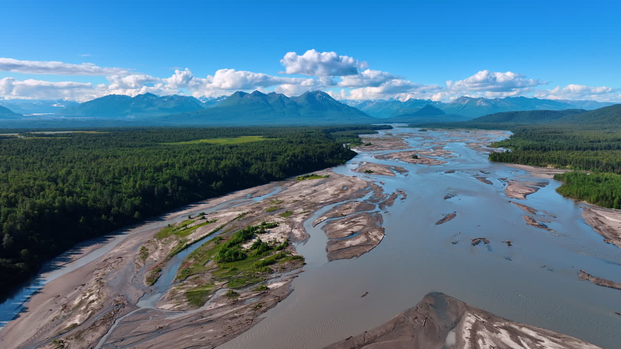 Sorvolando l'ampio fiume poco profondo nel fitto bosco. Montagne incredibili sotto le nuvole soffici sullo sfondo. Natura dell'Alaska ripresa con drone.
