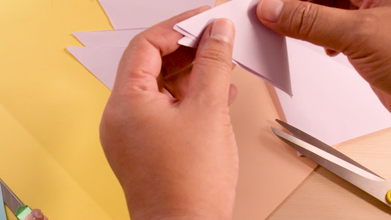 Primo piano di mani che piegano con precisione un foglio rosa su un'area di lavoro gialla, con delle forbici nelle vicinanze