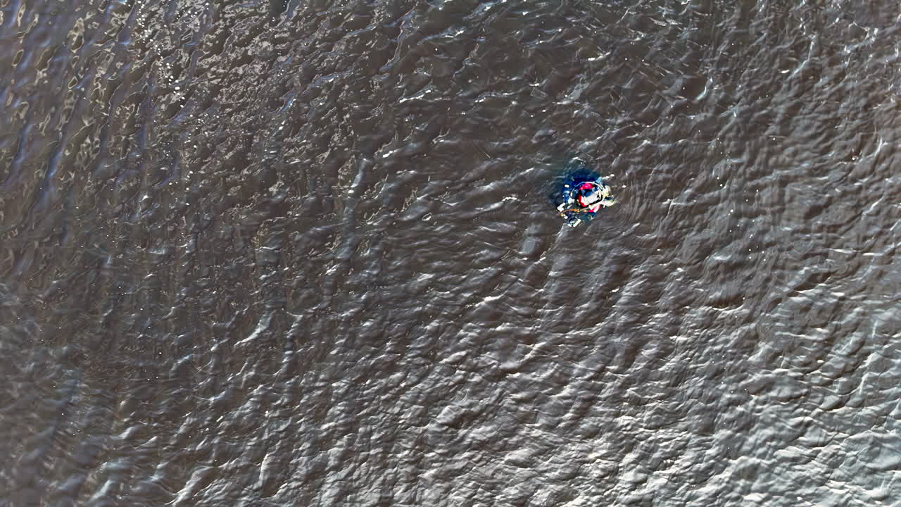 A single diver glides across textured ripples beneath a steady overhead drone, bright gear punctuating the gray surface and conveying training, vigilance, and quiet endurance in open water