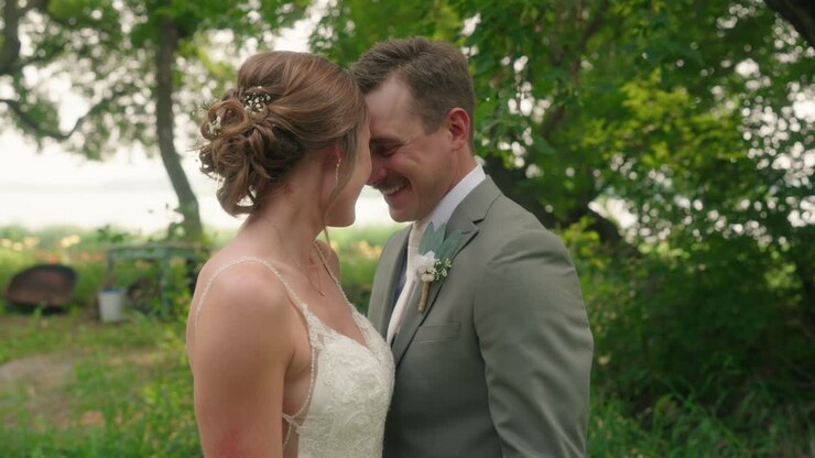 Bride and groom touch foreheads and smile in close-up with lake and sun rays in background