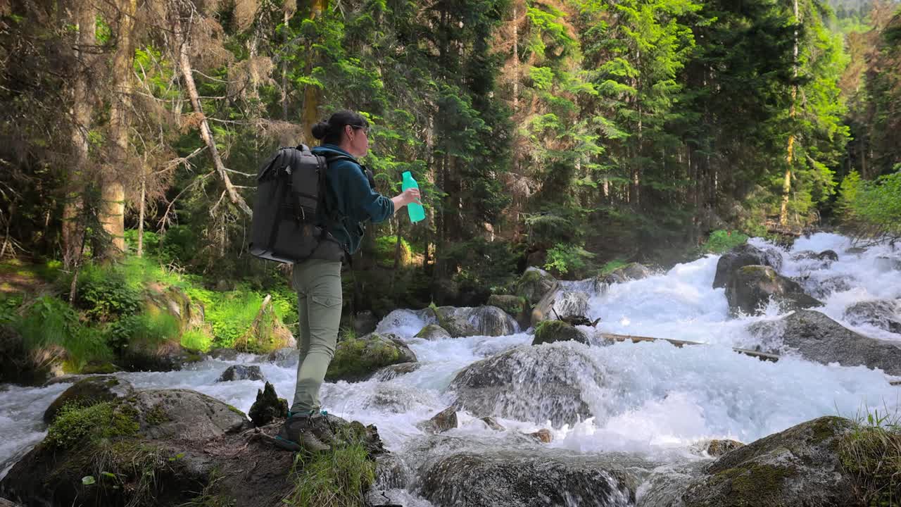 viajera con una mochila, bebiendo agua en la naturaleza en el bosque cerca de un río de montaña.