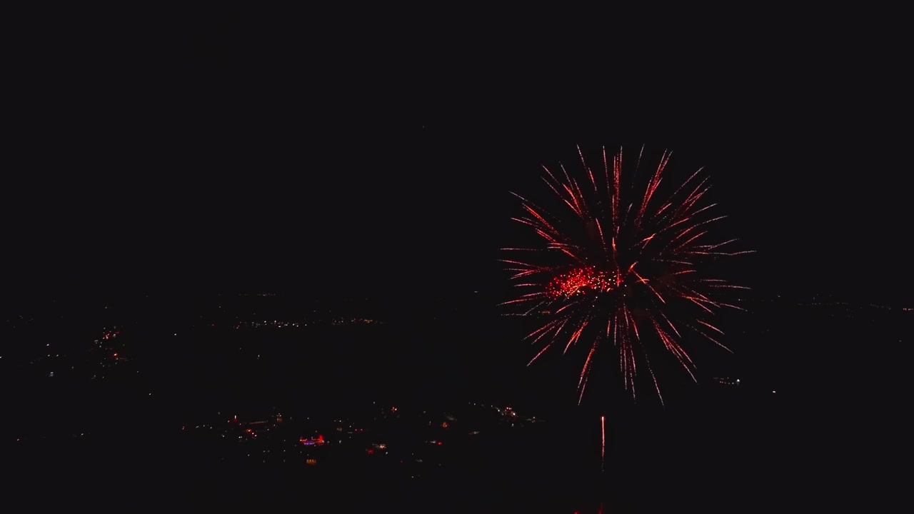 Bright fireworks burst in the night sky above the city. Families gather to watch the show. The event happens during a holiday celebration, creating a festive atmosphere.