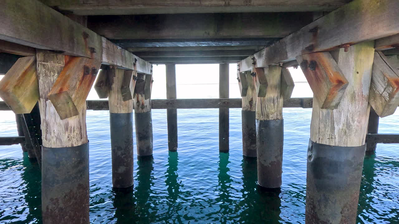 A close-up view of the underside of a jetty pier, showcasing the wooden support beams and the sturdy pylons submerged in the water