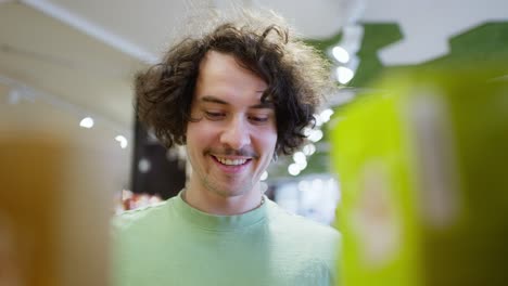 Nahaufnahme-Eines-Glücklichen-Brünetten-Kerls-Mit-Lockigem-Haar-In-Einem-Grünen-T-Shirt,-Der-Auf-Der-Theke-In-Einem-Supermarkt-Die-Roten-Waren-Auswählt,-Die-Er-Braucht