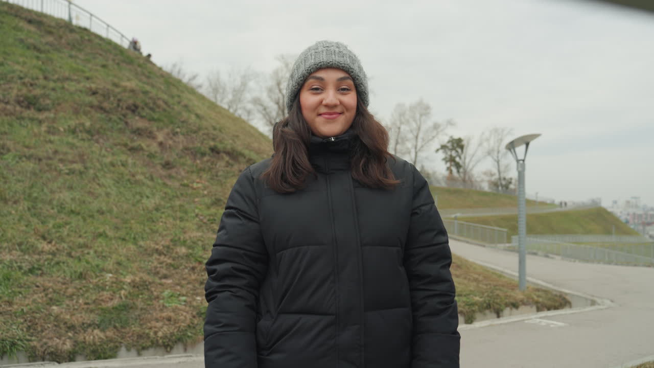 Una ballerina del parco posa dietro una ringhiera. La donna, con berretto di lana, irradia un sorriso sereno, indossando un piumino. Un sentiero aperto e una collina fanno da sfondo, con una coreografia sottile e una posa sicura, in un'atmosfera invernale.