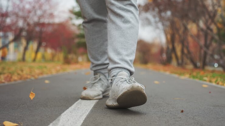 Close up leg view of athlete walking on tarred road as dry foliage sways with wind, autumn leaves scattered along path with soft blur background of trees in park setting on cool breezy day