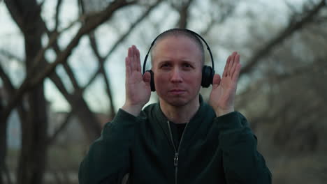 a man in a green jacket and headphones frames his face with his hands in a forest, his focused expression and distinctive pose stand out against the serene wooded background