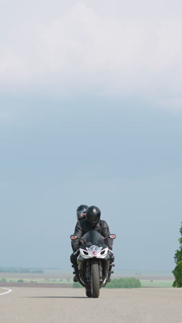 cyclist with passenger rides modern motorcycle along highway against cloudy sky and trees at countryside on sunny day low angle shot slow motion