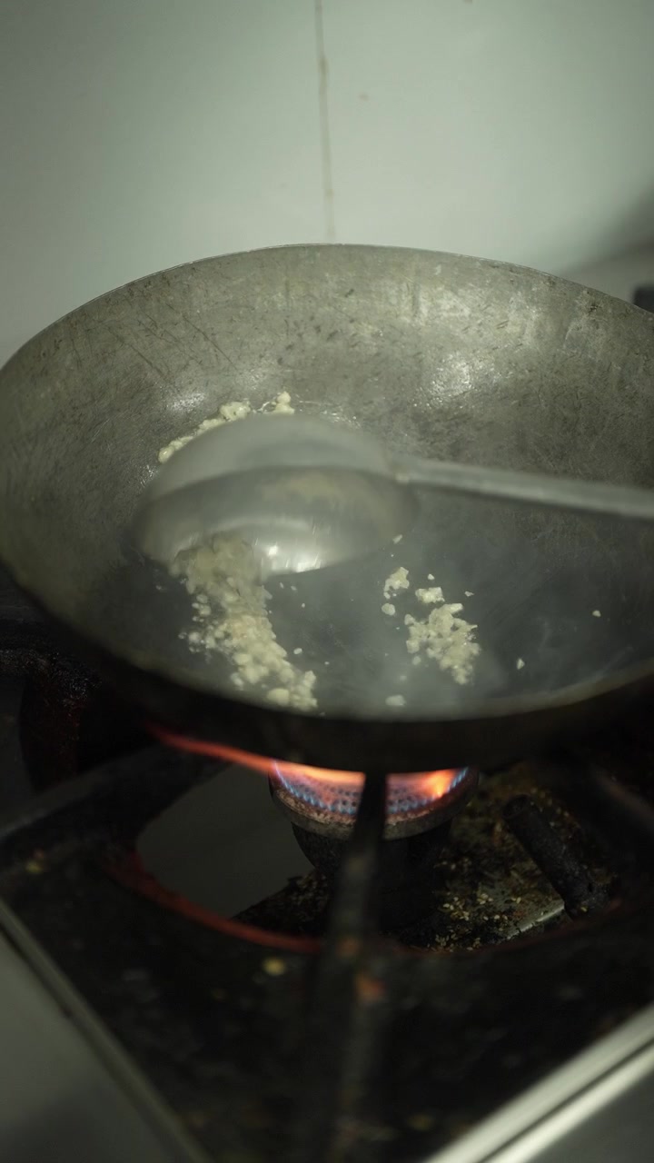 Riprese verticali ravvicinate di aglio tritato aggiunto e saltato in burro fuso in un wok di ferro caldo su una fiamma a gas, con un mestolo che mescola l'aglio