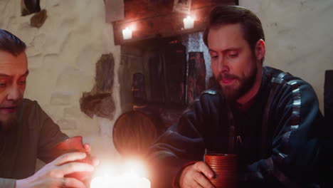 un hombre golpea la mesa con el puño cerca de un amigo en una taberna medieval. un tipo enojado se queja de la vida y bebe alcohol con otro en una vieja posada. un invitado emocional descansa en el bar.