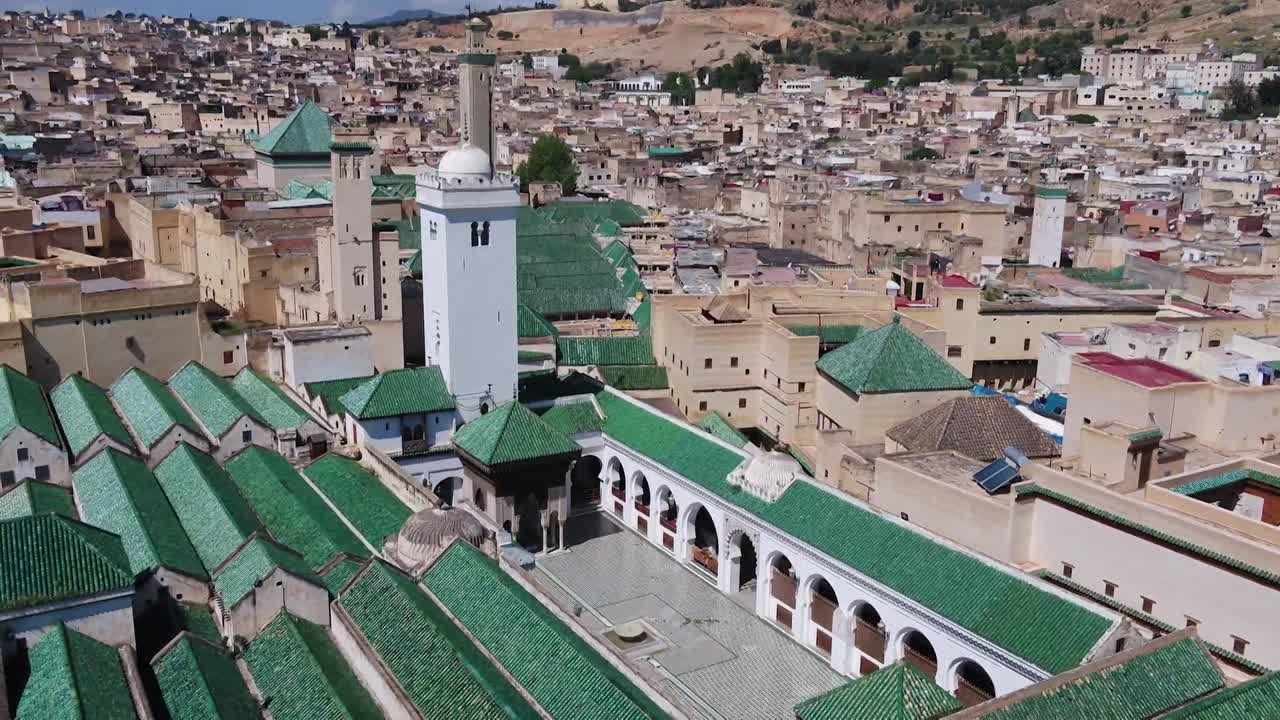 Bela imagem de drone focando nos telhados de azulejos verdes geométricos de Al-Qarawiyyin. Apresenta os minaretes brancos históricos e a arquitetura tradicional circundante da medina de Fez El Bali