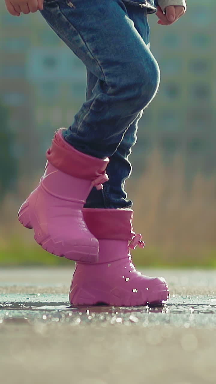 slow motion close up kid in wet pants and rubber boots jumps on puddle stomping his foot in park after rain in sunny summer weather