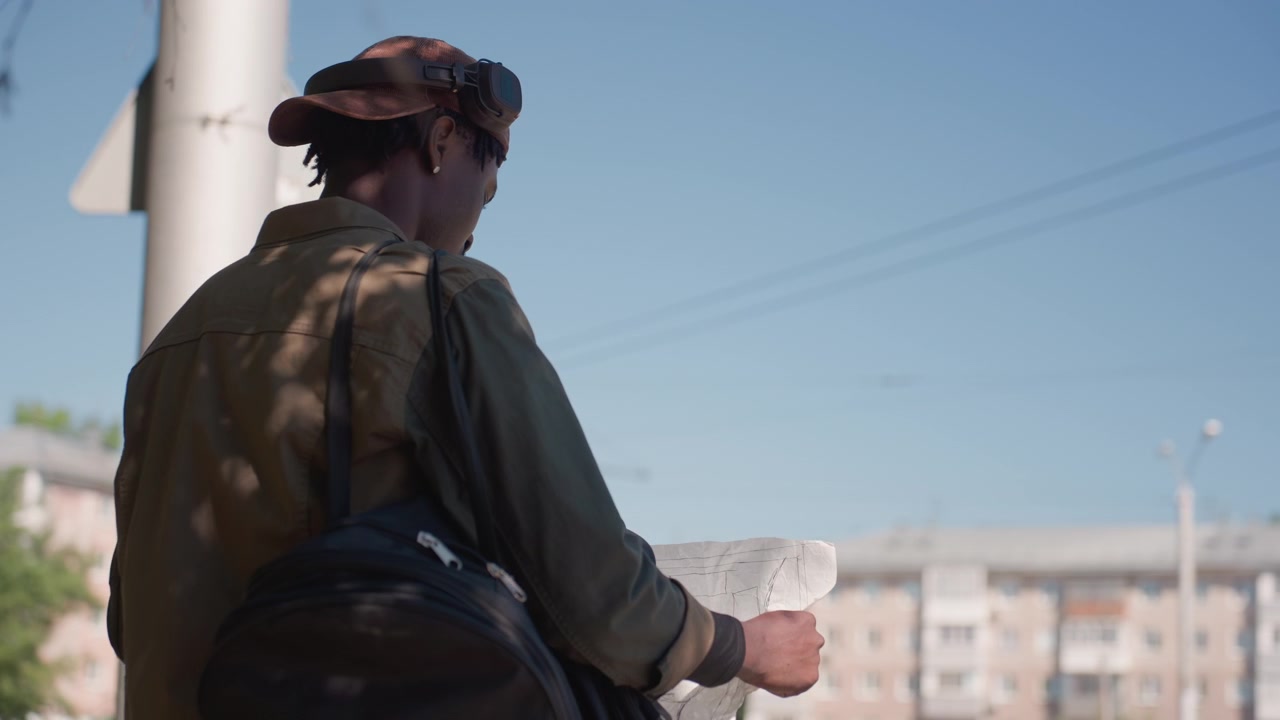 Primo piano di un uomo che esamina dei documenti all'aperto con cielo sereno e sfondo urbano. L'uomo tiene in mano i documenti mentre indossa un berretto, una giacca e uno zaino; la scena mostra luce diurna, ombre tenui ed edifici distanti.