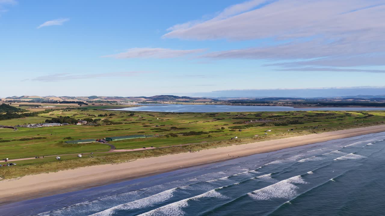 Drönare glider över sandstrand, golfbana och vågor under starkt dagsljus vid St Andrews
