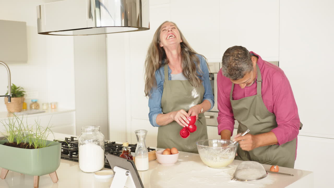 Cooking, multiracial senior couple laughing and mixing ingredients in modern kitchen, at home