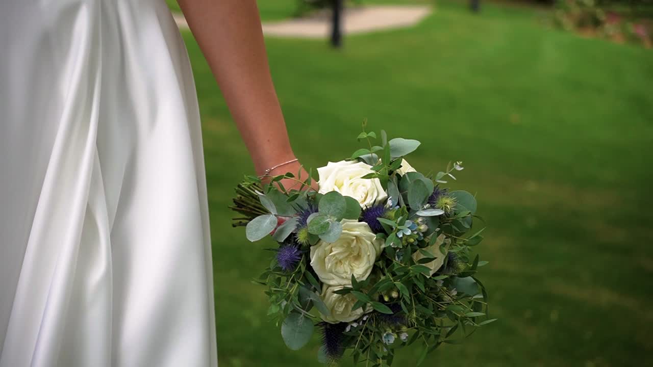 Primo piano di una sposa che cammina all'aperto nel parco tenendo in mano un semplice bouquet da sposa di rose bianche e verde con sfondo sfocato