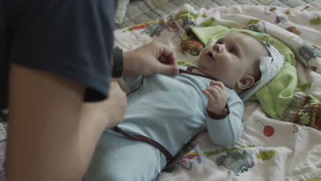 young mother finishing dressing up her baby boy infant, who lies on the coachwith colorful bedsheet