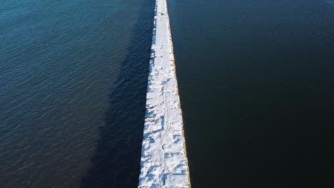 Reveladora-Vista-Aérea-Del-Muelle-De-Hormigón-Cubierto-De-Nieve-Y-Hielo-En-El-Tranquilo-Mar-Báltico,-Puerto-De-Liepaja-En-Un-Soleado-Día-De-Invierno,-Disparo-De-Drones-De-Gran-Angular-Lento-Que-Avanza-La-Cámara-Inclinada-Hacia-Arriba