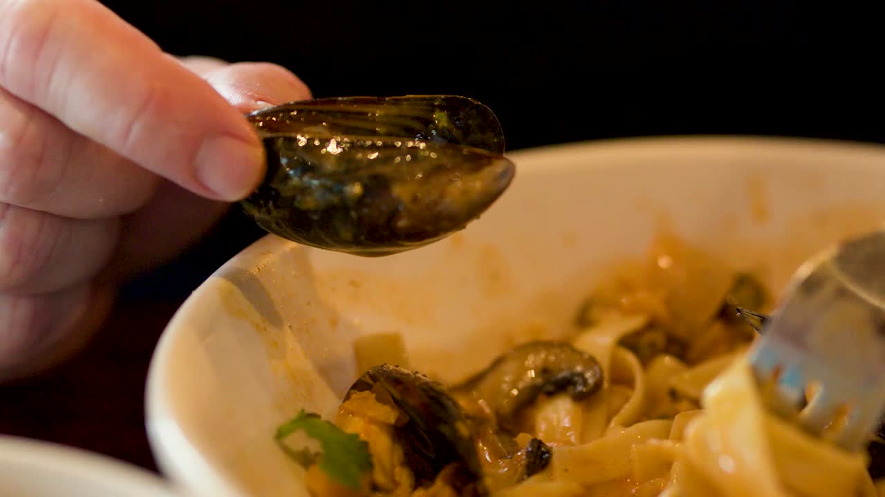 Una mano tiene in mano un guscio di cozza mentre una forchetta arrotola dei cremosi noodles in una ciotola
