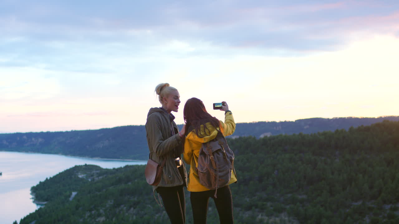 dos mujeres tomando una selfie en una pintoresca cima de la montaña con vistas a un lago al atardecer