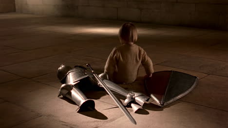 a child sits beside a suit of knight's armor and weapons