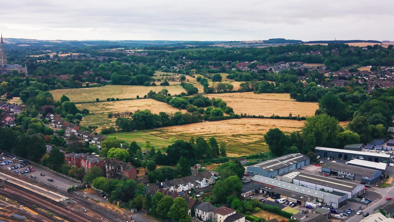 Riprese aeree con drone dei prati acquatici di Salisbury in estate, con campi dorati, case a schiera, una linea ferroviaria e il bordo della cattedrale visibile