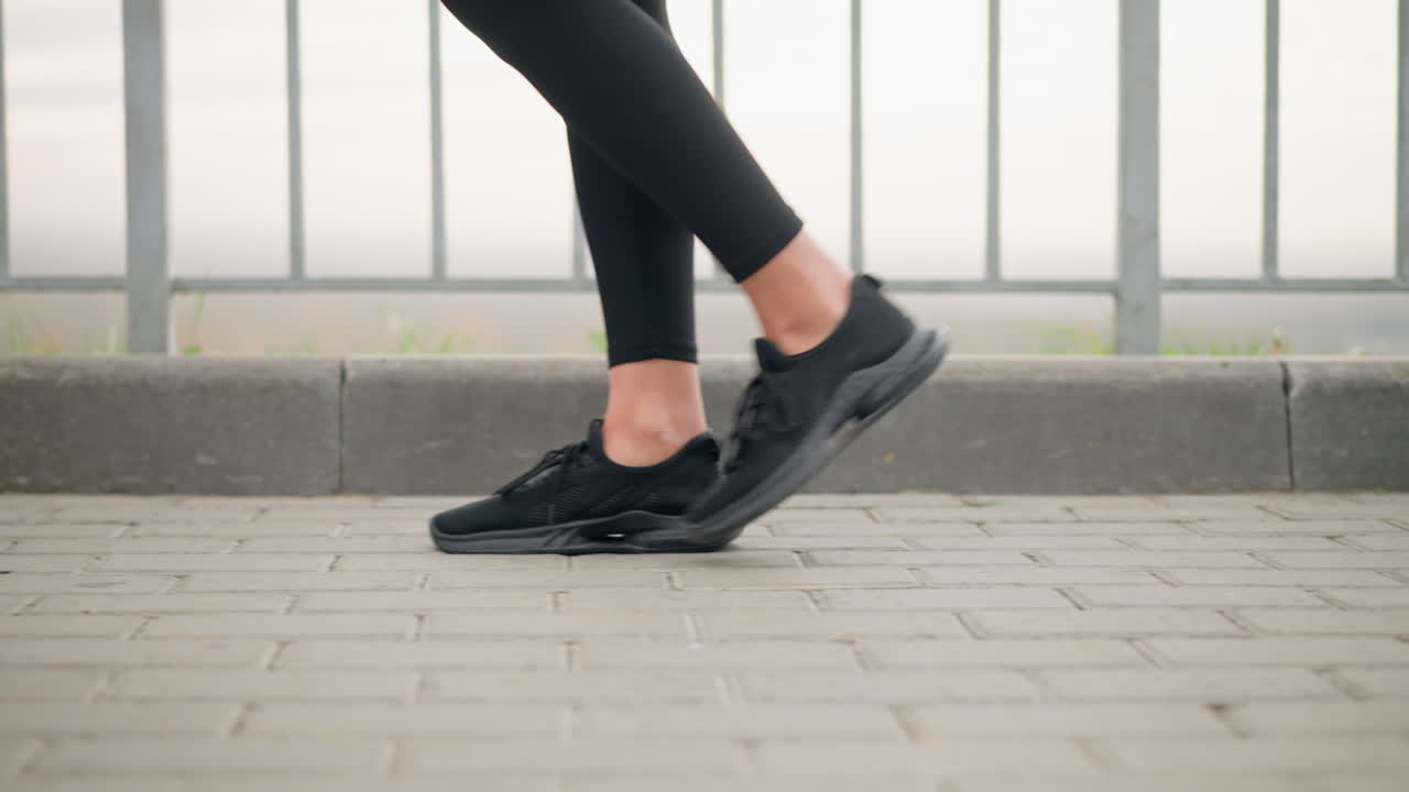 Leg view of woman in black sneakers and black leggings walking confidently on interlocked pavement beside iron rail, showcasing a casual, relaxed outdoor stroll in urban environment
