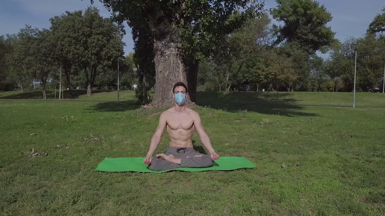 Jovem com máscara praticando yoga no parque no tapete verde, relaxamento em pose de lótus.