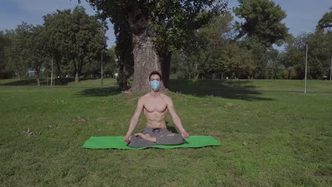 Young-man-with-the-mask-doing-yoga-in-the-park-on-the-green-mat,-lotus-pose-relaxation