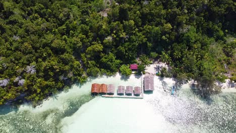 aerial drone rising above a row of waterfront beach huts overlooking ocean on rainforest covered island in raja ampat, west papua, indonesia