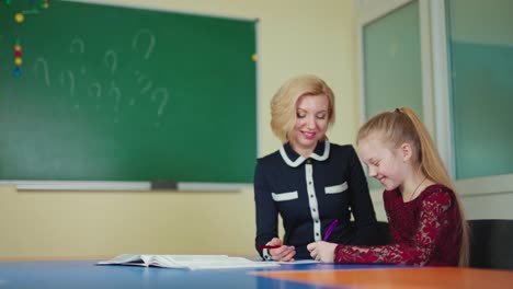 positive teacher with a cute schoolgirl. little student learning writing with a tutor. experienced teacher helps studying elementary pupil at school table.