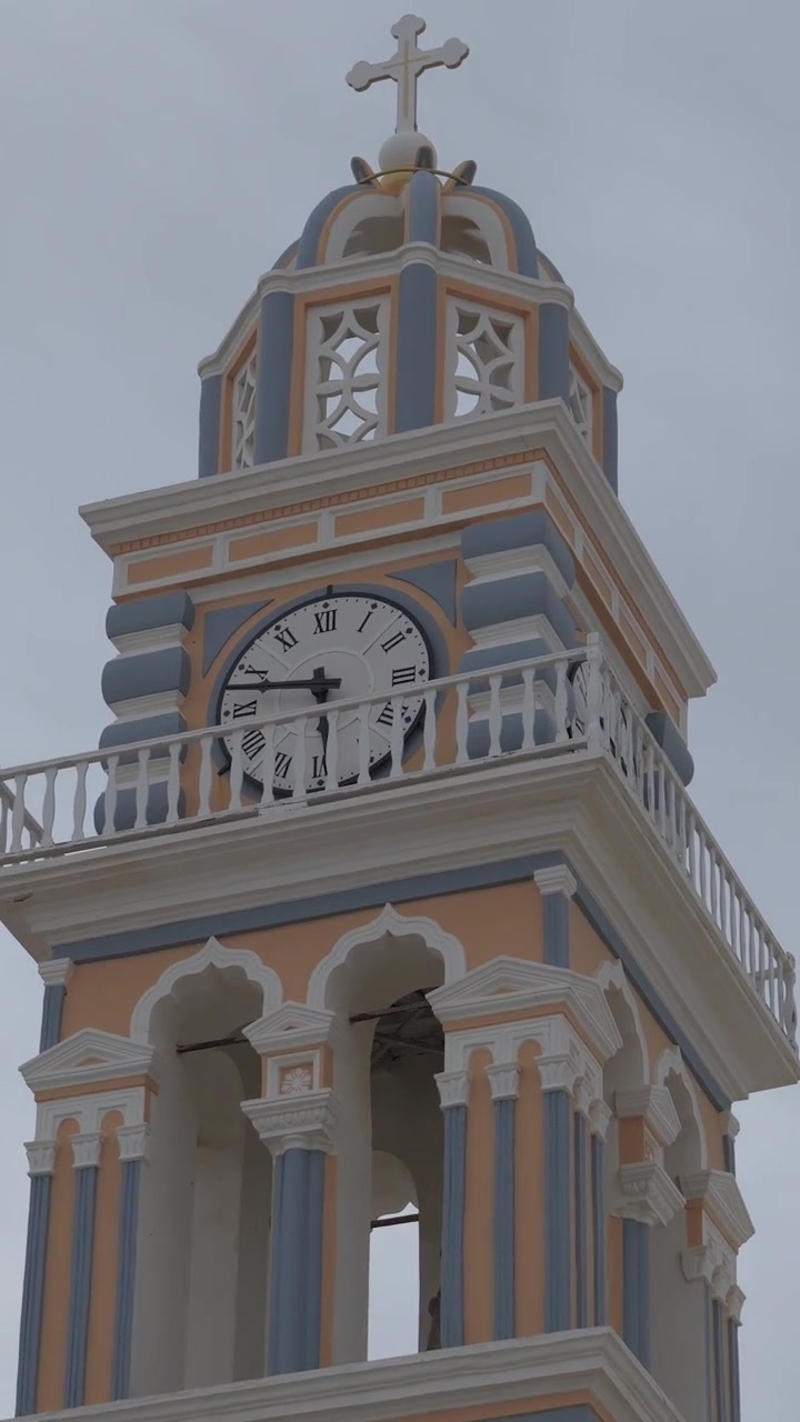 Vertical tilt up to the clock tower of the Holy Cathedral of Saint John the Baptist in Fira, Santorini, highlighting its historic Cycladic architecture.