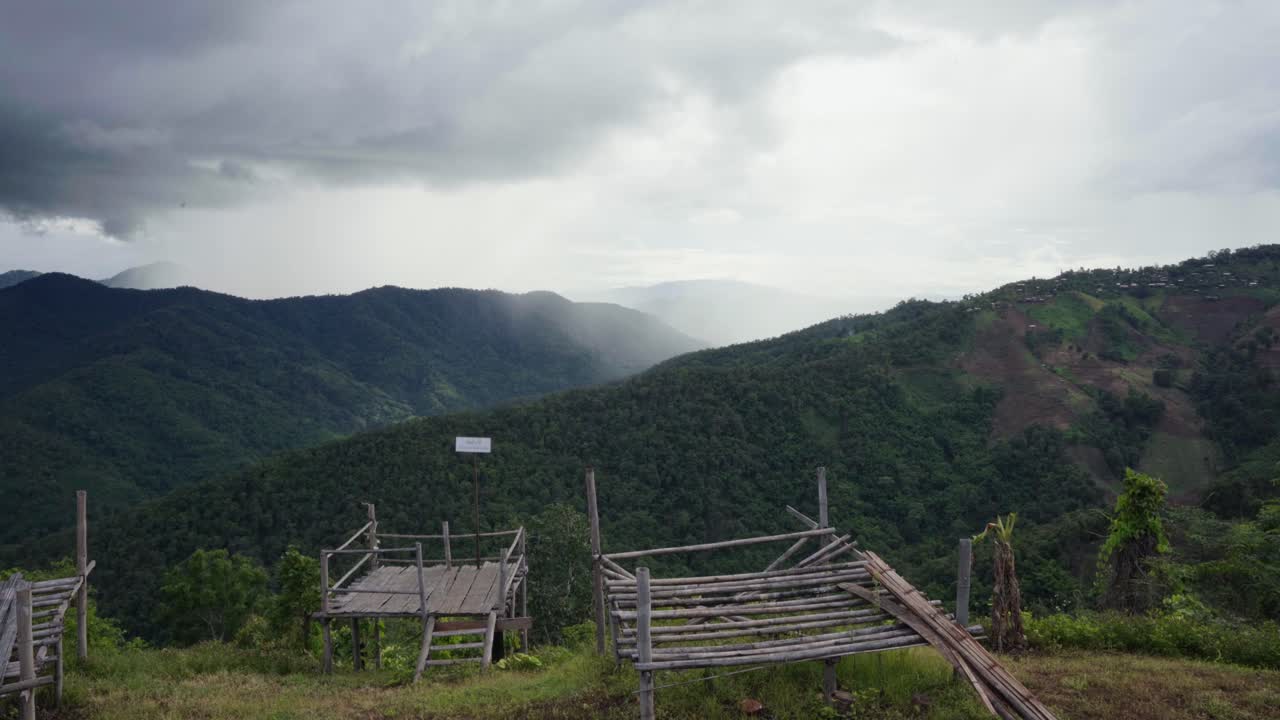 desde una cubierta de observación de bambú iluminada por el sol con un panel de observación en la cima de una montaña en mae tang, tailandia, las nubes de lluvia oscuras cubren un valle lejano que avanza hacia un pueblo iluminado que pronto lloverá.