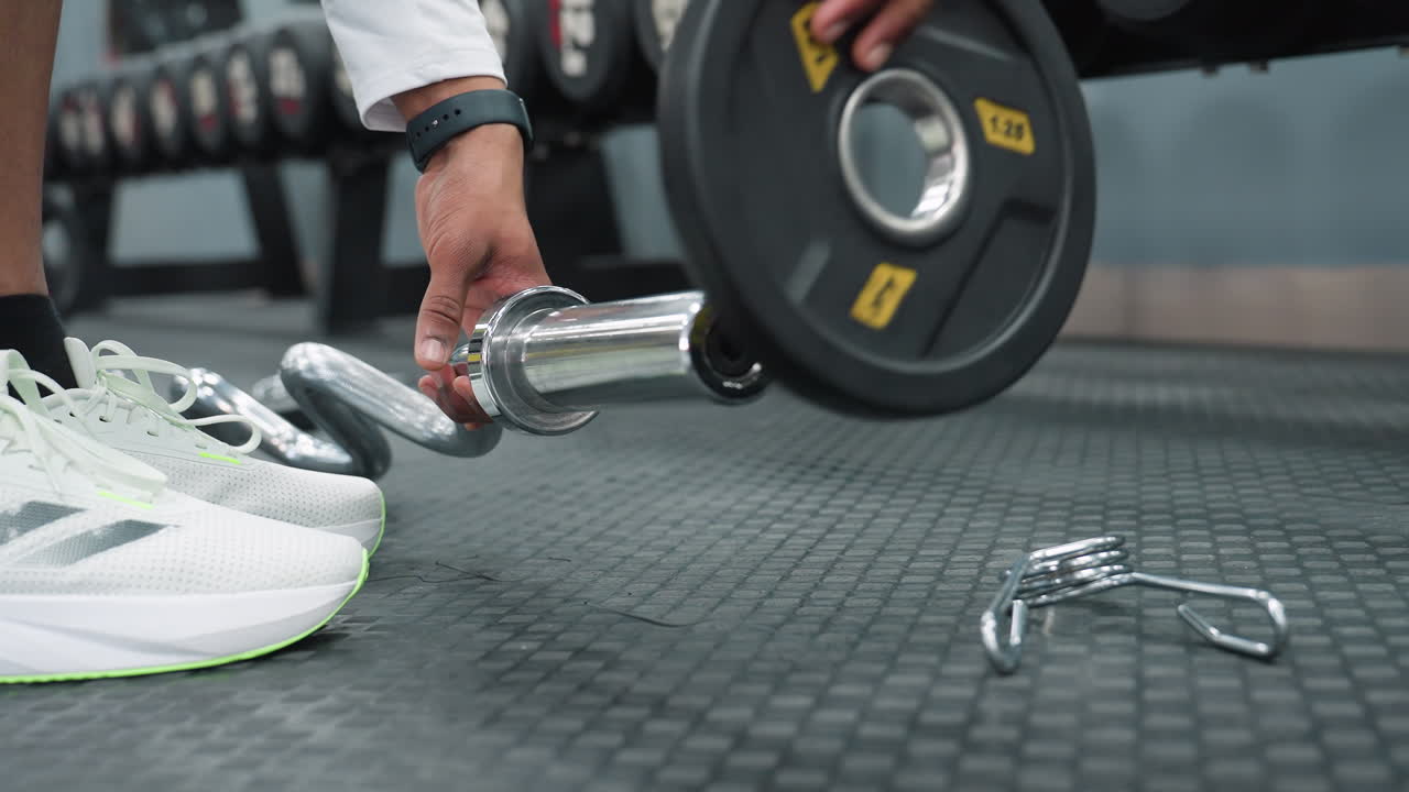 Close up hands of person securing barbell with metal spring clip on gym floor while getting ready for workout, wearing white sportswear and sneakers, with weights and equipment in background