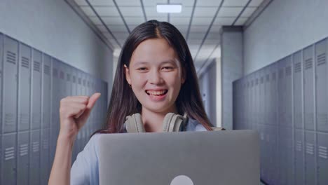 primer plano de una estudiante adolescente asiática con una mochila usando una computadora portátil y celebrando mientras está de pie en el pasillo