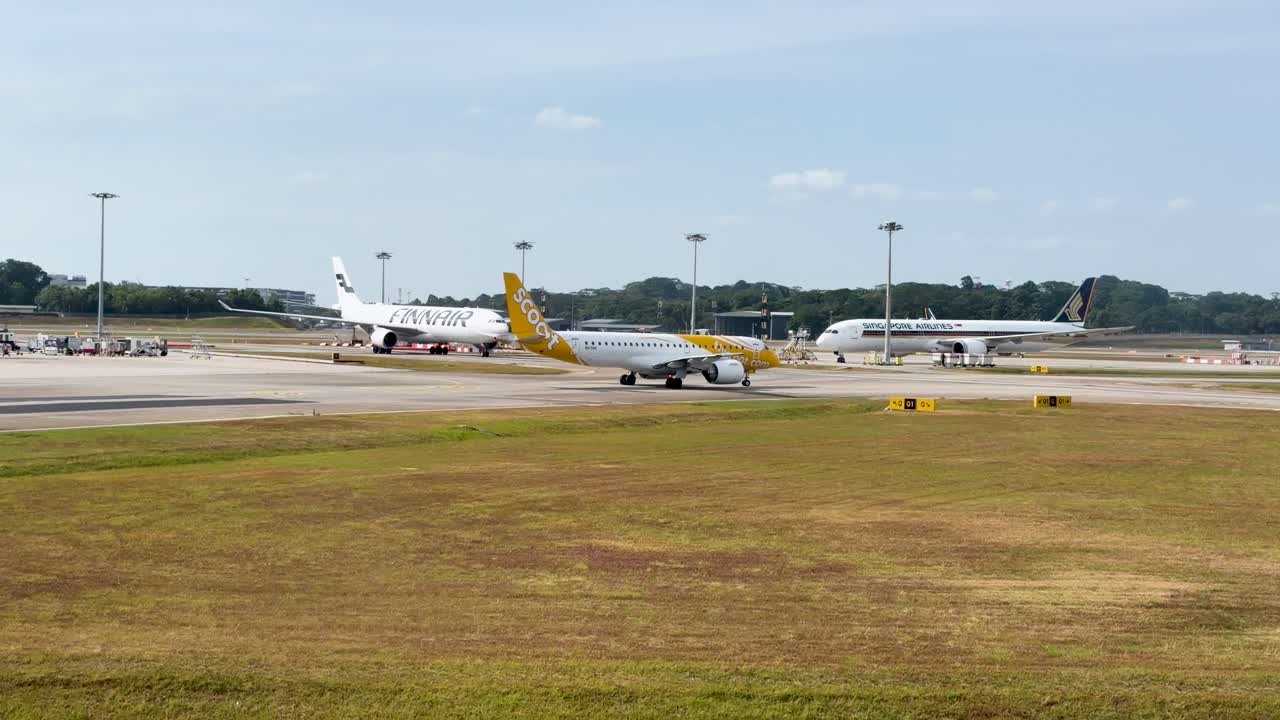 Ripresa grandangolare di numerosi aerei a reazione commerciali in rullaggio su una pista aeroportuale durante le ore diurne.